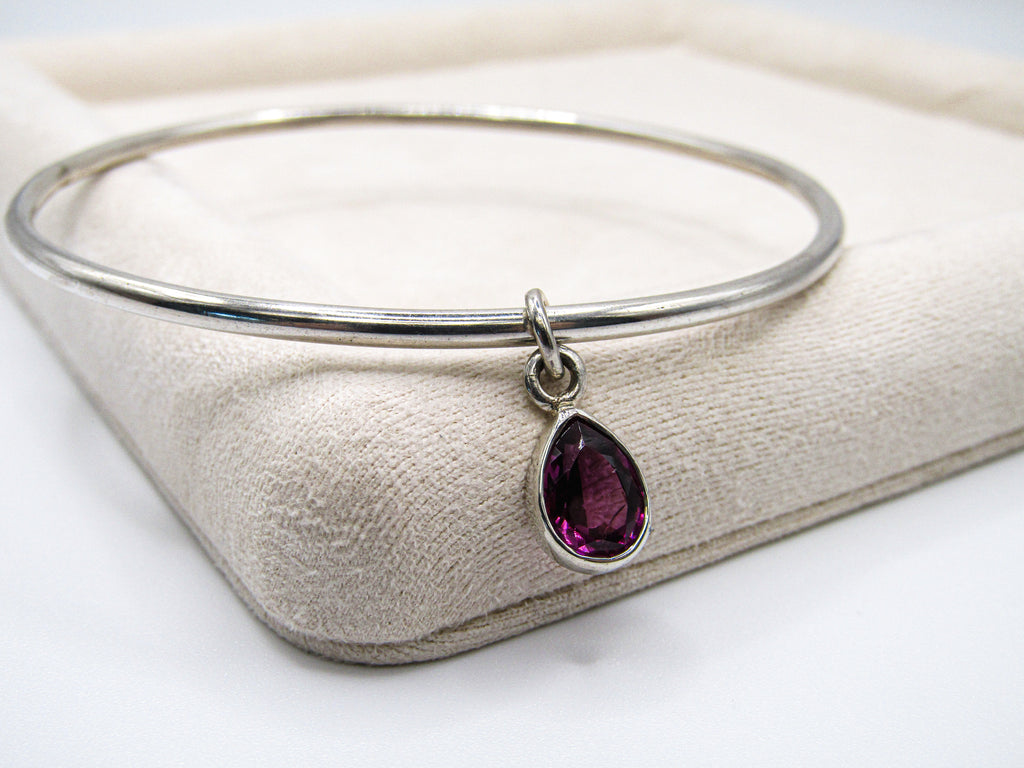 Silver bangle with a teardrop-shaped purple gemstone charm on a white background