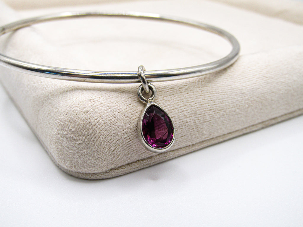 Silver bangle with a purple teardrop pendant on a beige fabric background