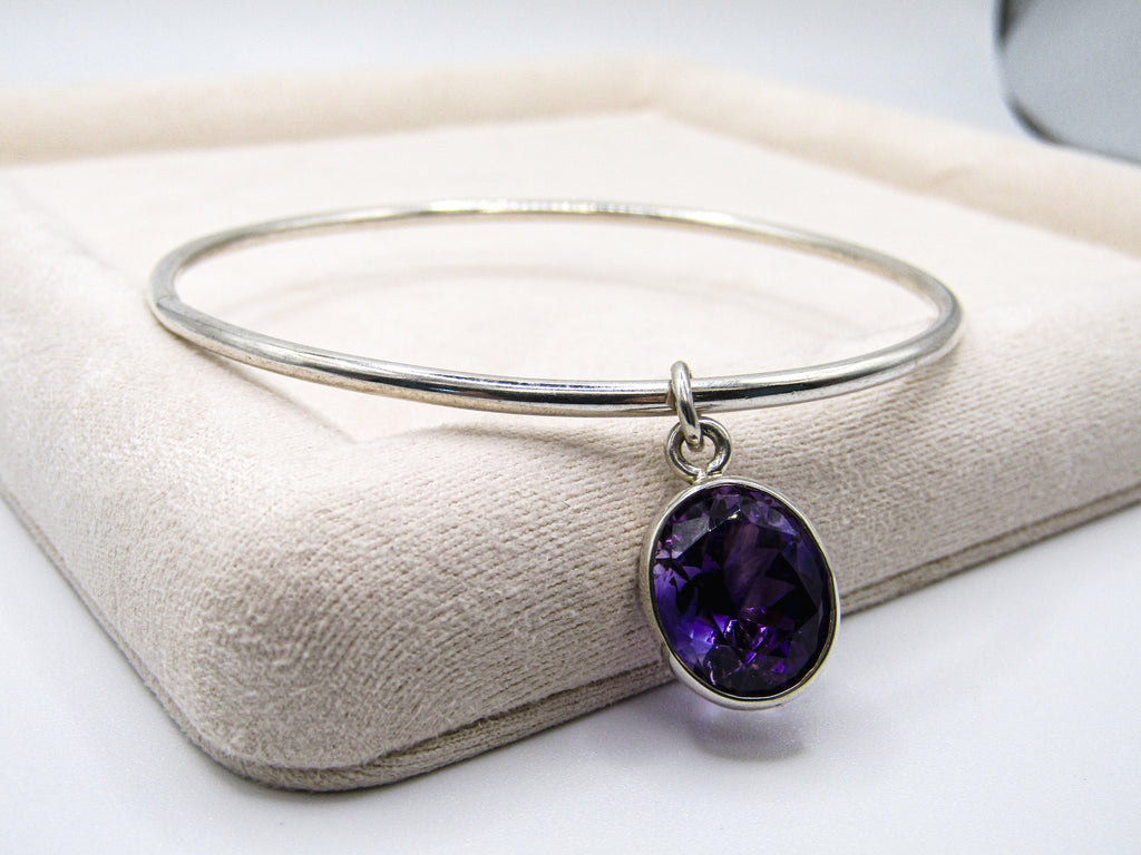 Silver bangle with a purple gemstone pendant on a beige cushion