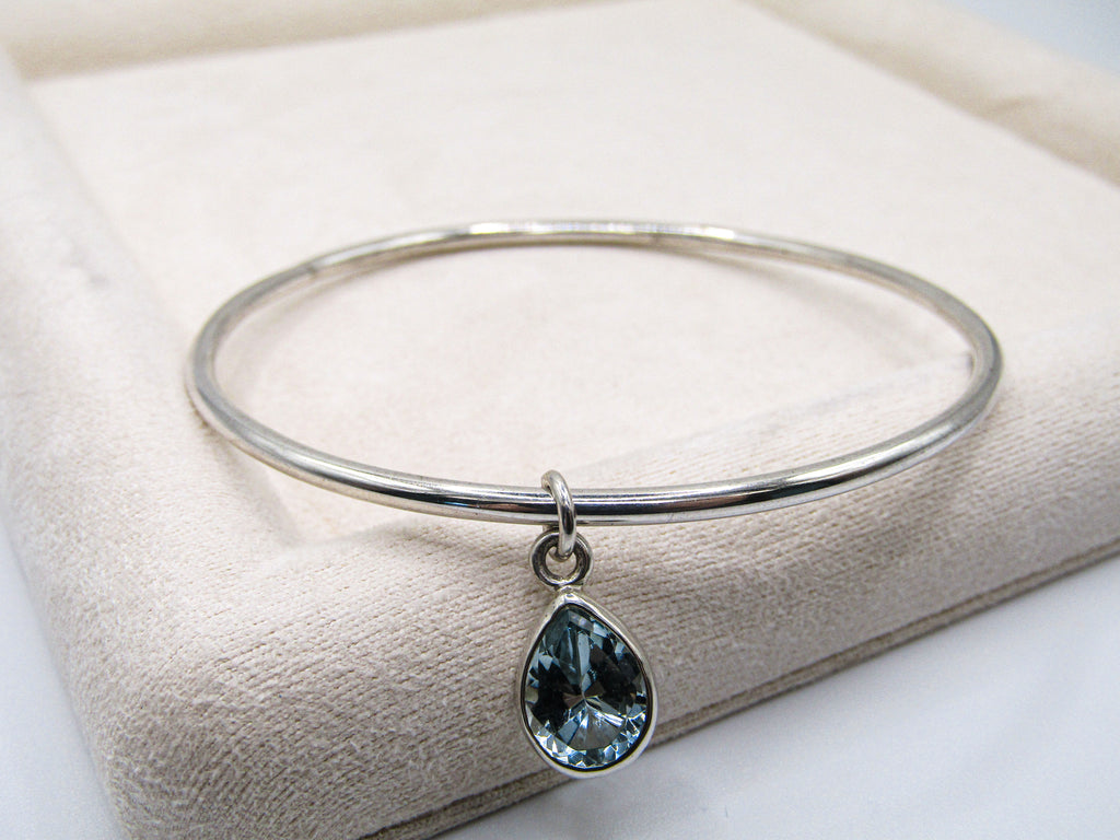 Silver bangle with a teardrop-shaped blue gemstone charm on a beige cushion.