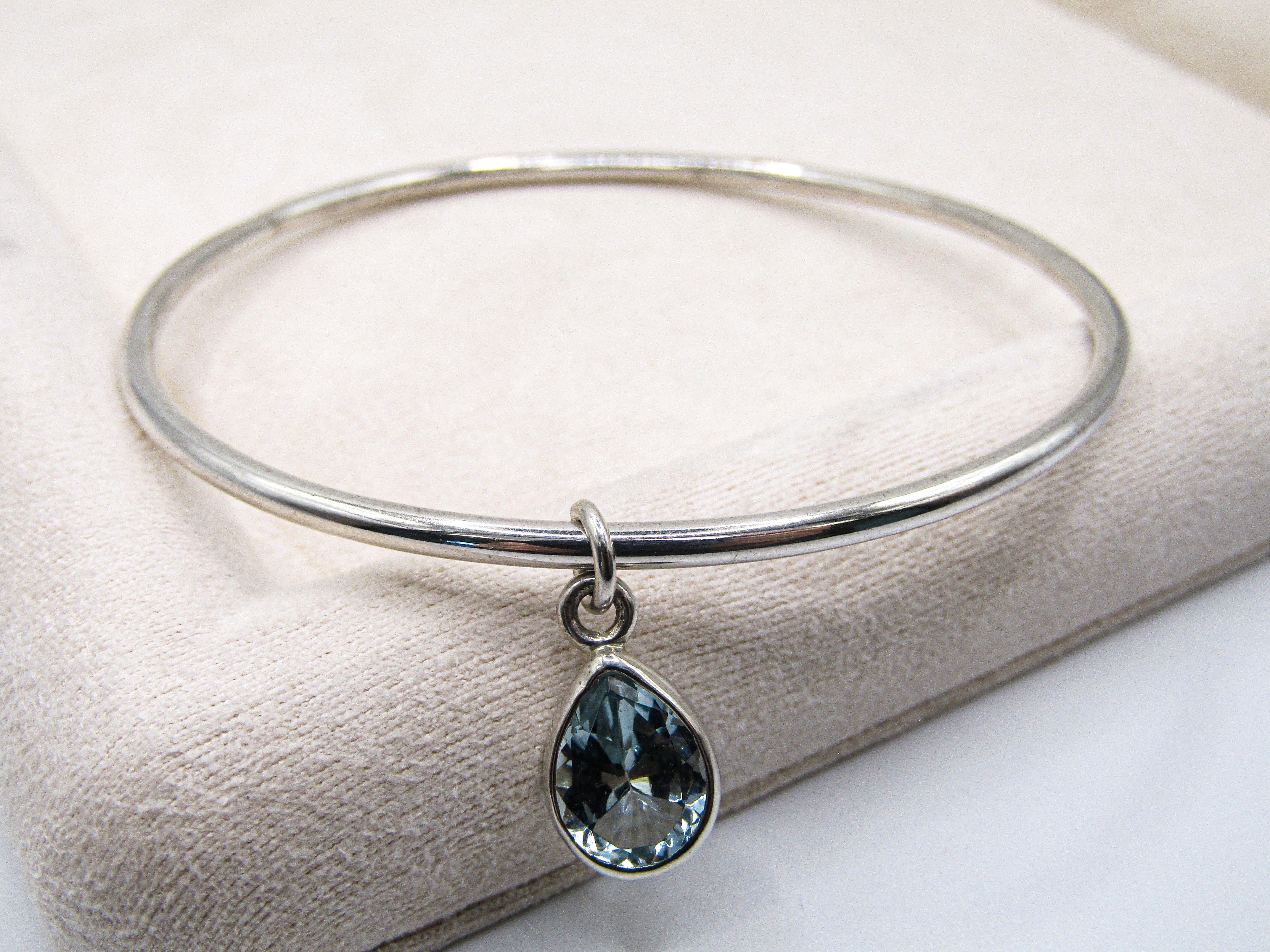 Silver bangle with a teardrop-shaped blue gemstone charm on a white surface
