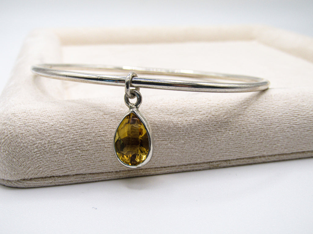 Silver bangle with a yellow teardrop pendant on a textured beige surface.