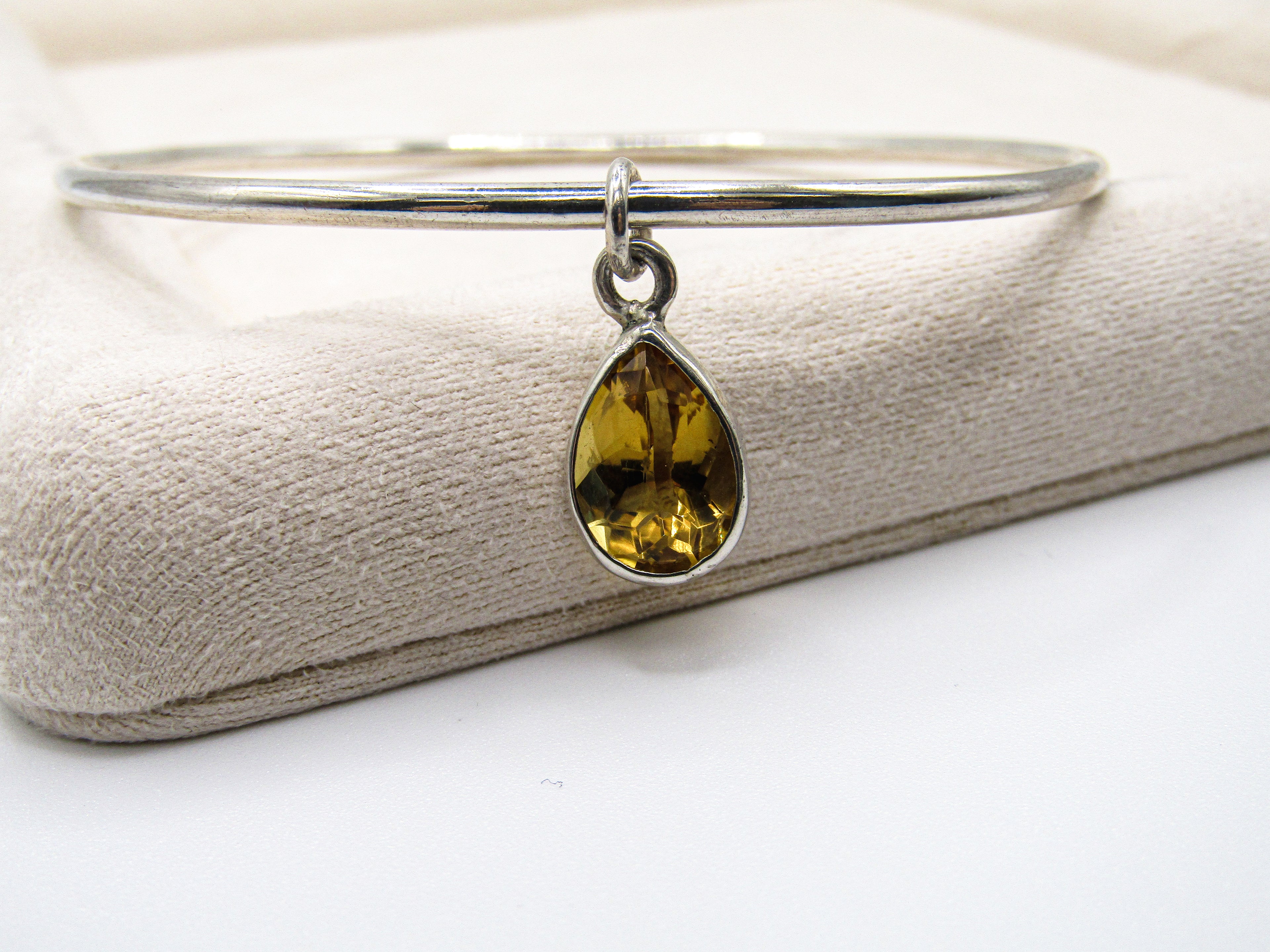 Silver bangle with a teardrop-shaped yellow gemstone on a textured beige surface.