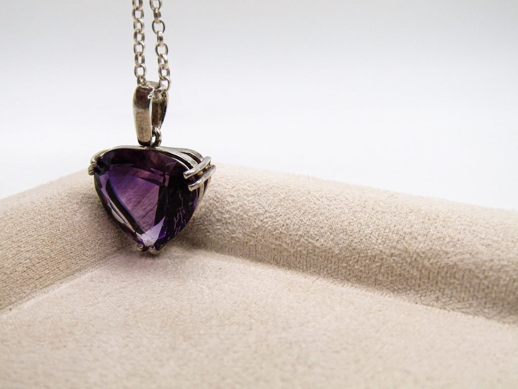 Purple heart-shaped gemstone pendant on a chain, displayed on a beige cushion with a white background.