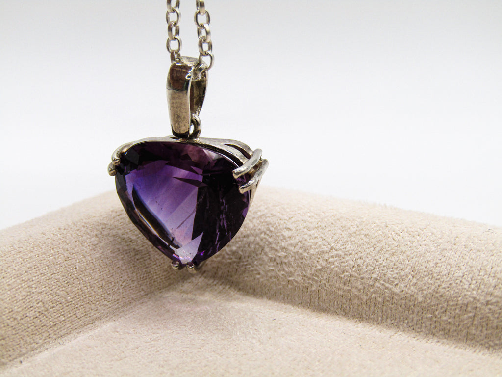 Purple heart-shaped gemstone pendant on a chain, displayed on a beige cushion with a white background.