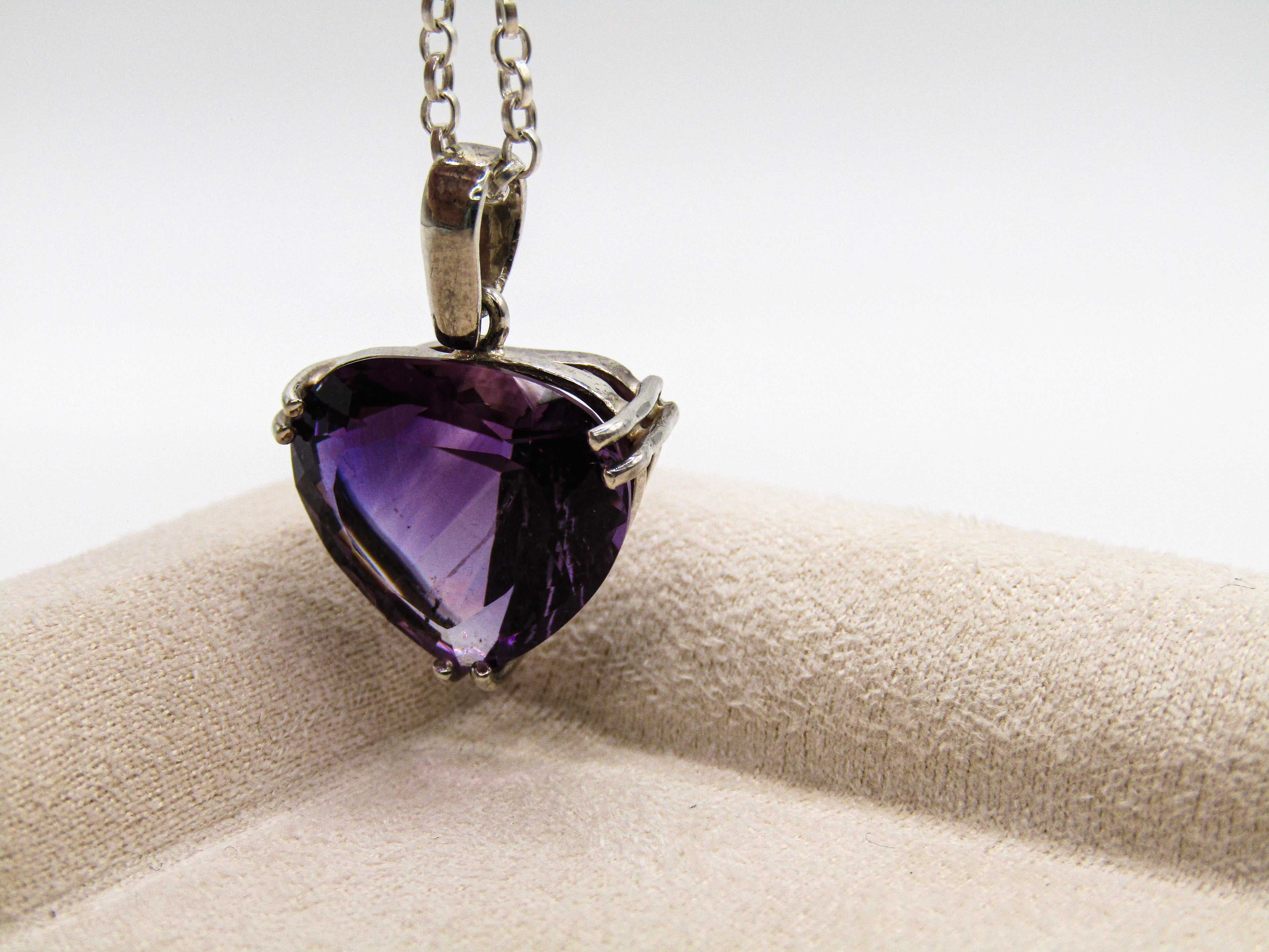Purple heart-shaped gemstone pendant on a chain, displayed on a beige cushion with a white background.